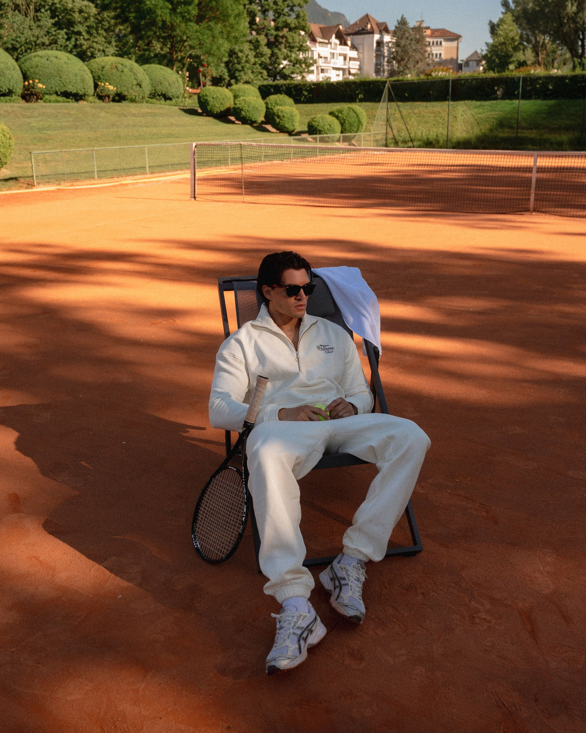 Model seated on a clay tennis court wearing cream Country Club sweatpants with a matching quarter-zip sweatshirt.