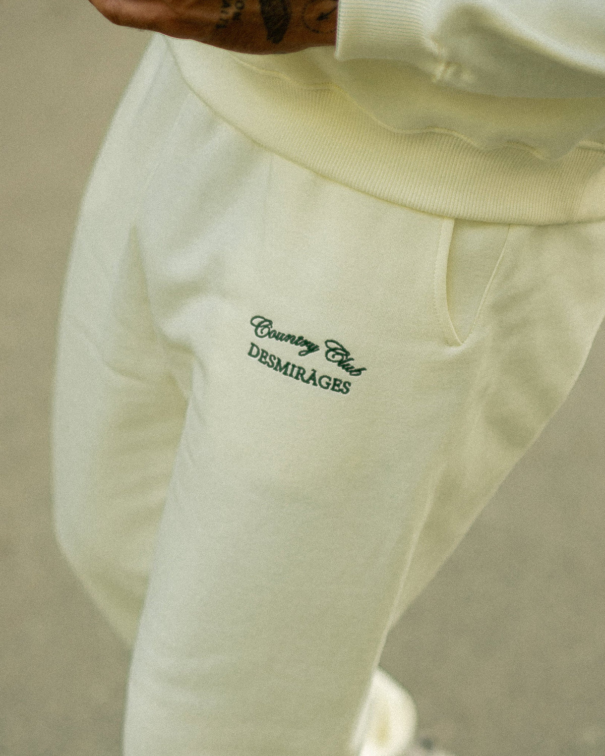Cream Country Club sweatpants worn with a matching sweatshirt, showing the green embroidered thigh logo and side pocket.