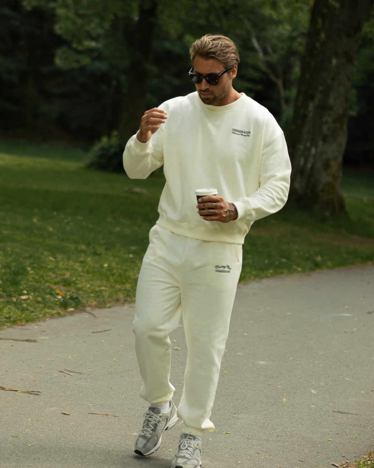 Model walking outdoors in cream Country Club sweatpants and a matching sweatshirt, wearing sunglasses and carrying a takeaway coffee.