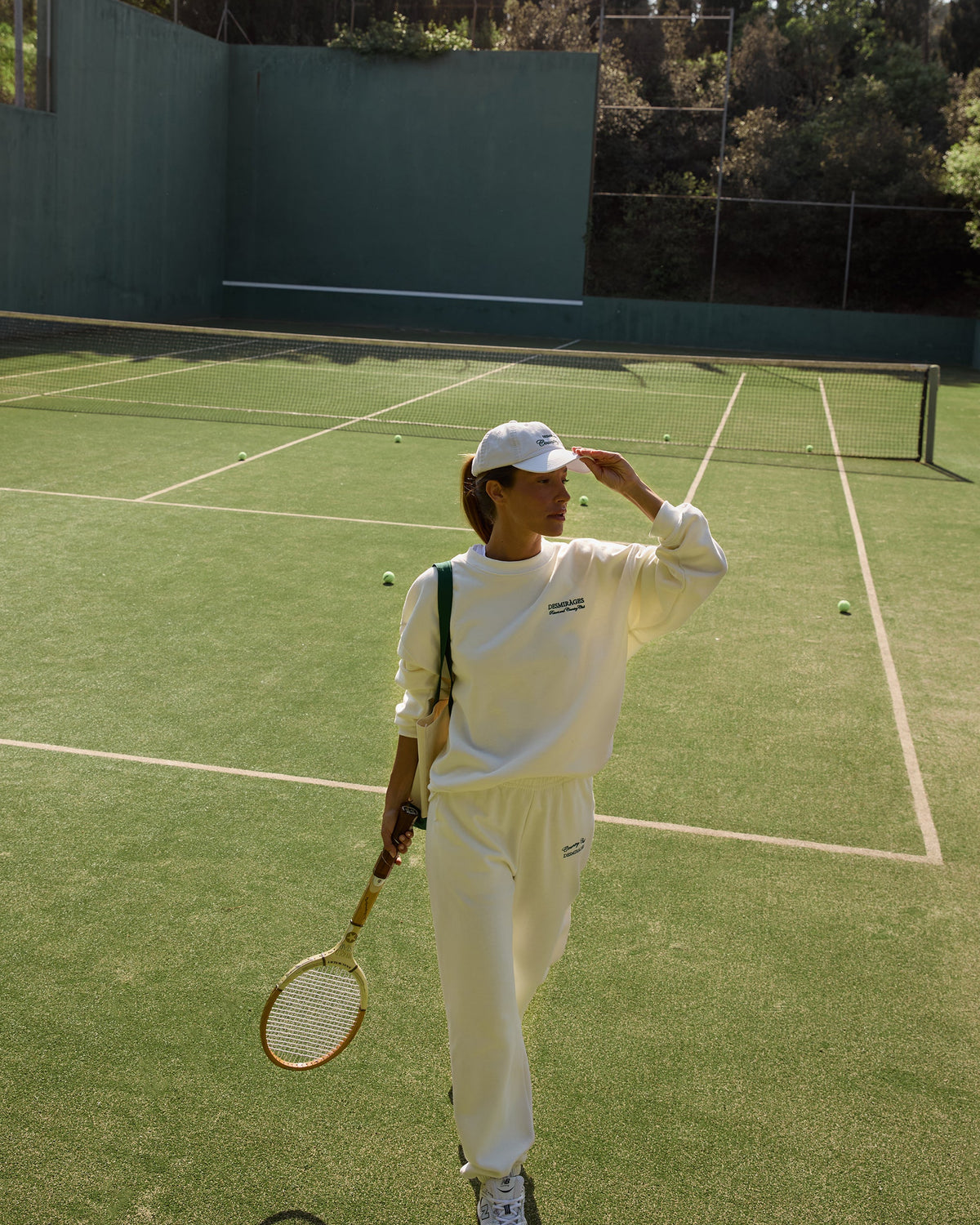 Model wearing cream Country Club sweatpants, a matching sweatshirt and white cap, holding a tennis racket on a green court.