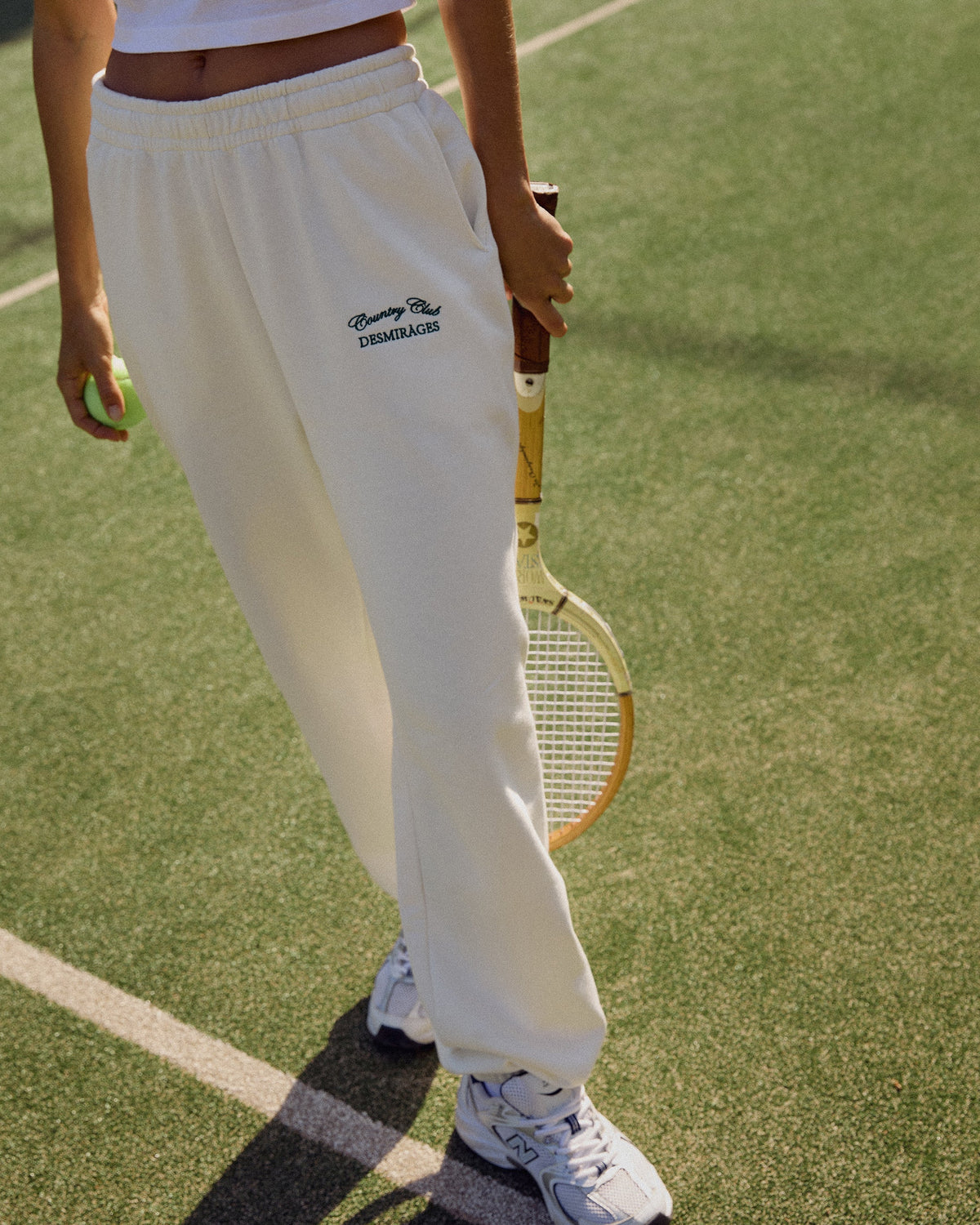 Model wearing cream Country Club sweatpants with a white cropped top and trainers, holding a racket on a tennis court.