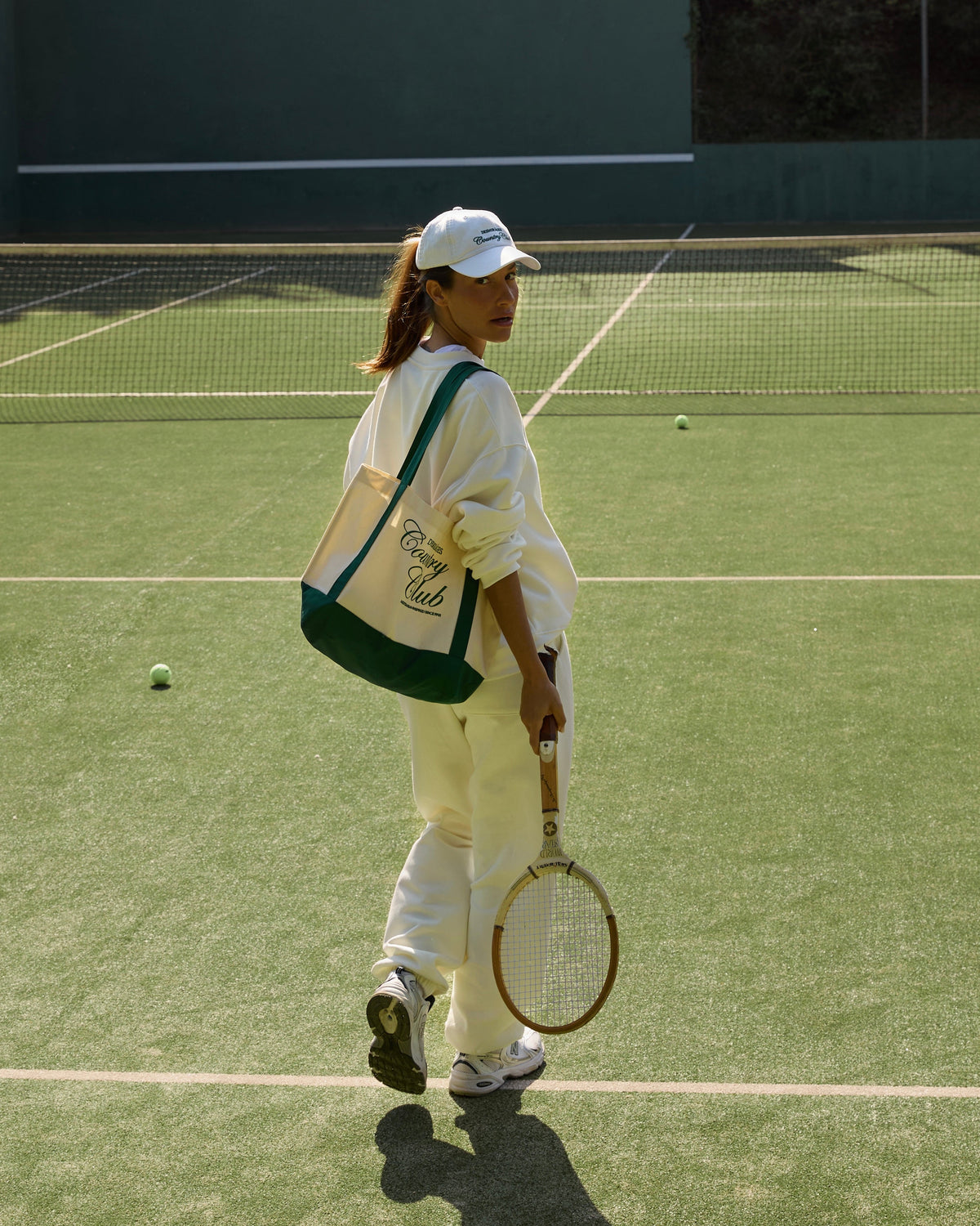 Model wearing cream Country Club sweatpants and a matching sweatshirt, carrying a green-trimmed tote bag and tennis racket on court.