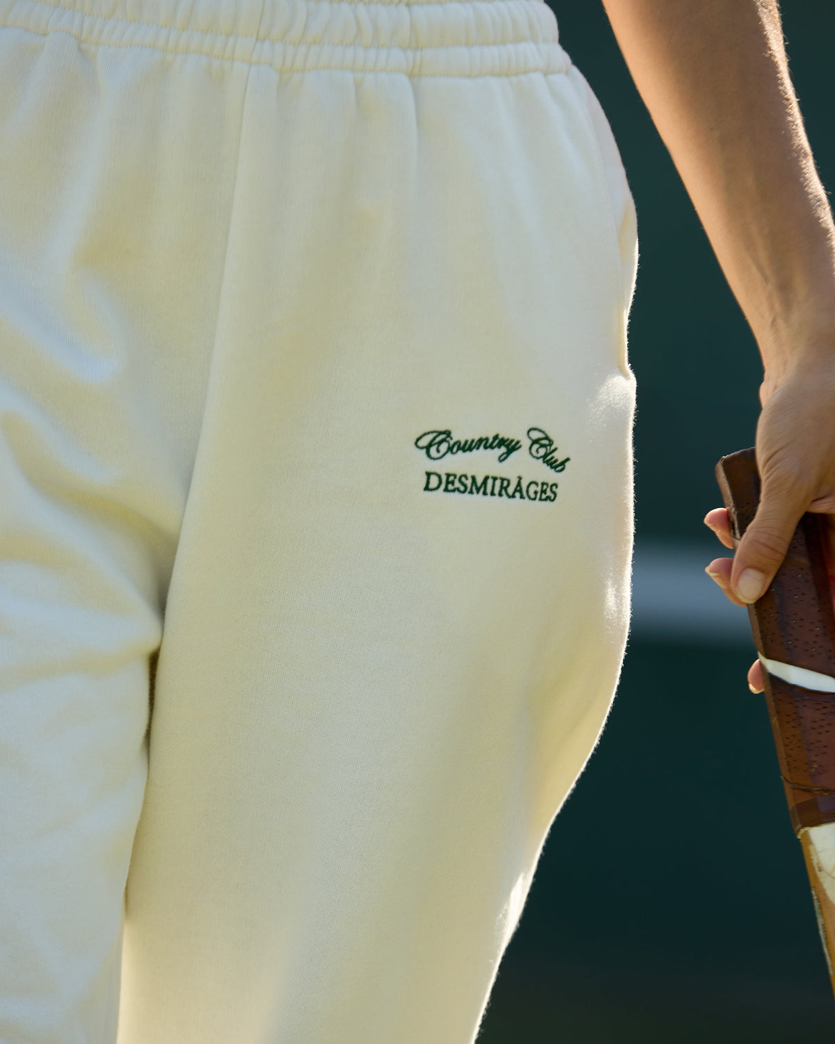 Close-up of the elastic waistband, side pocket and green DESMIRAGES Country Club embroidery on cream sweatpants.