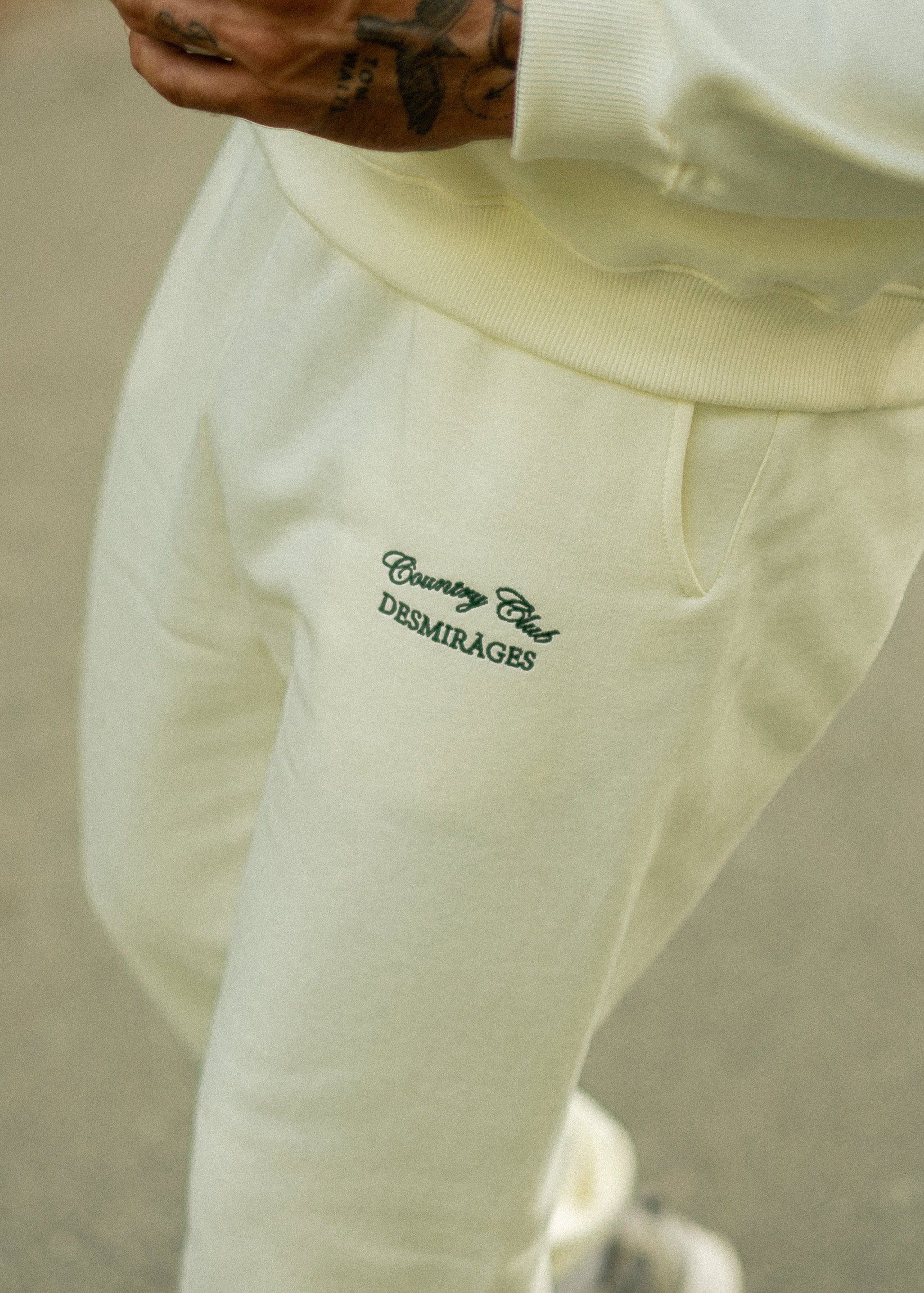 Cream Country Club sweatpants worn with a matching sweatshirt, showing the green embroidered thigh logo and side pocket.