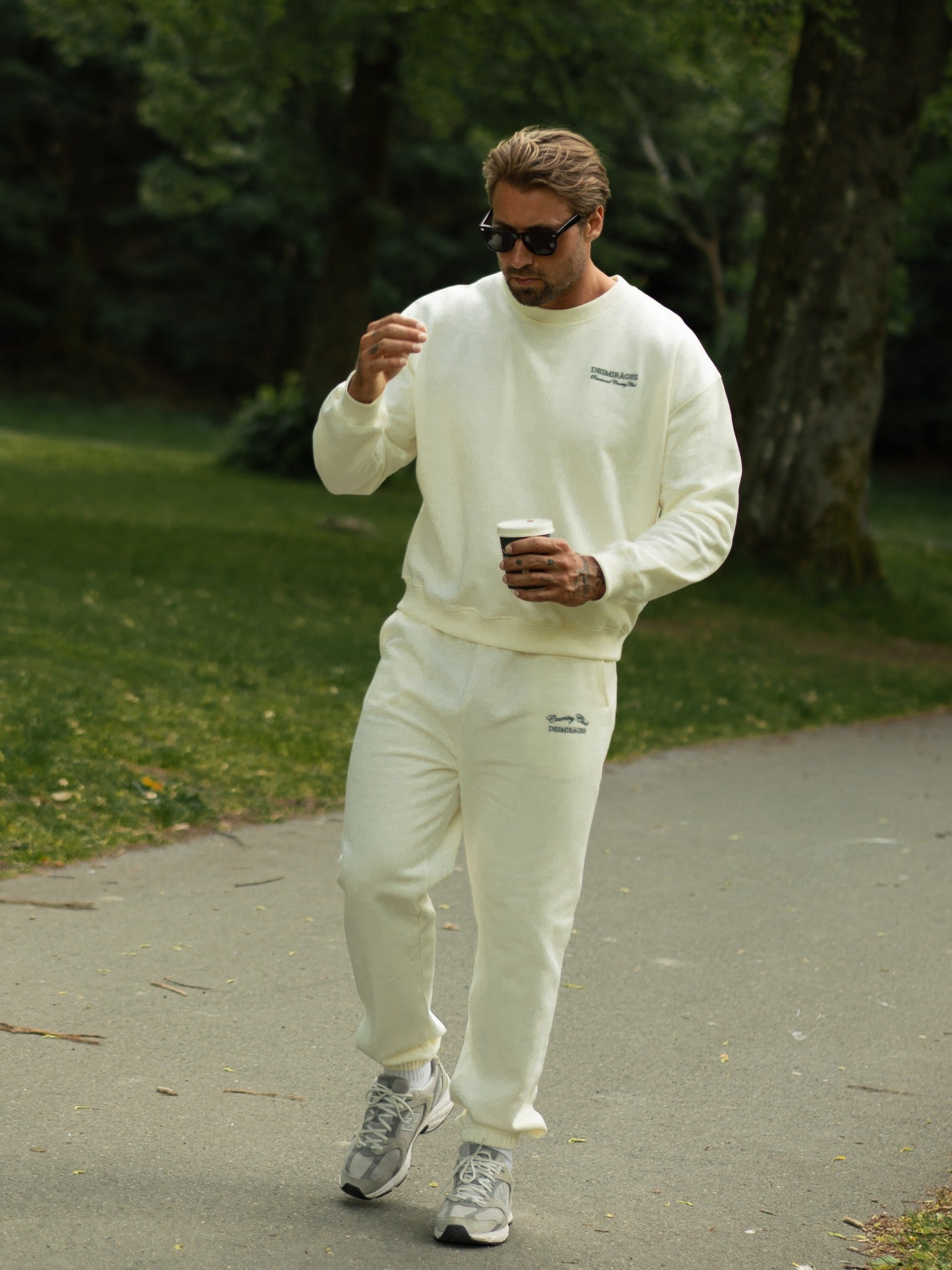 Model walking outdoors in cream Country Club sweatpants and a matching sweatshirt, wearing sunglasses and carrying a takeaway coffee.