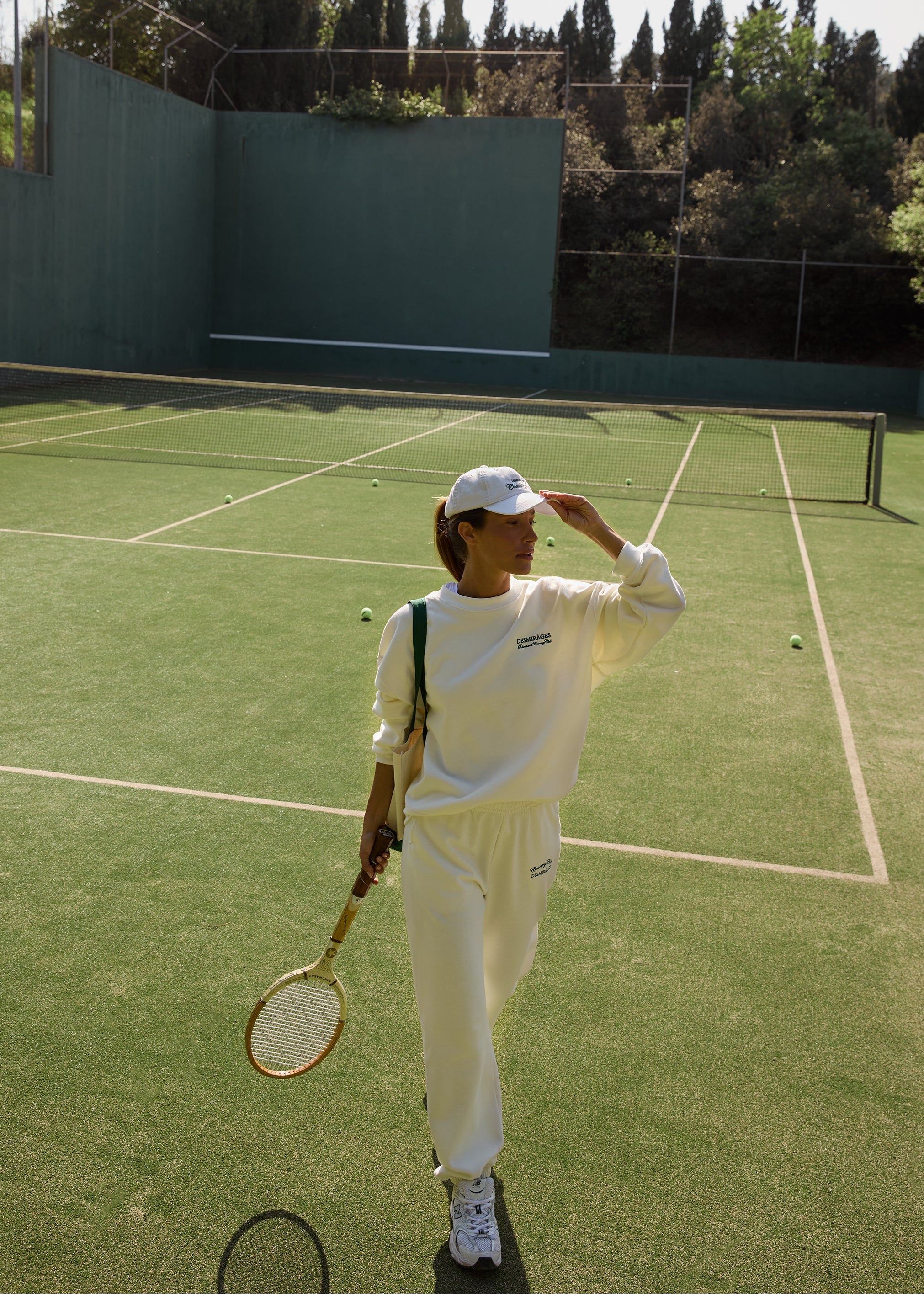 Model wearing cream Country Club sweatpants, a matching sweatshirt and white cap, holding a tennis racket on a green court.