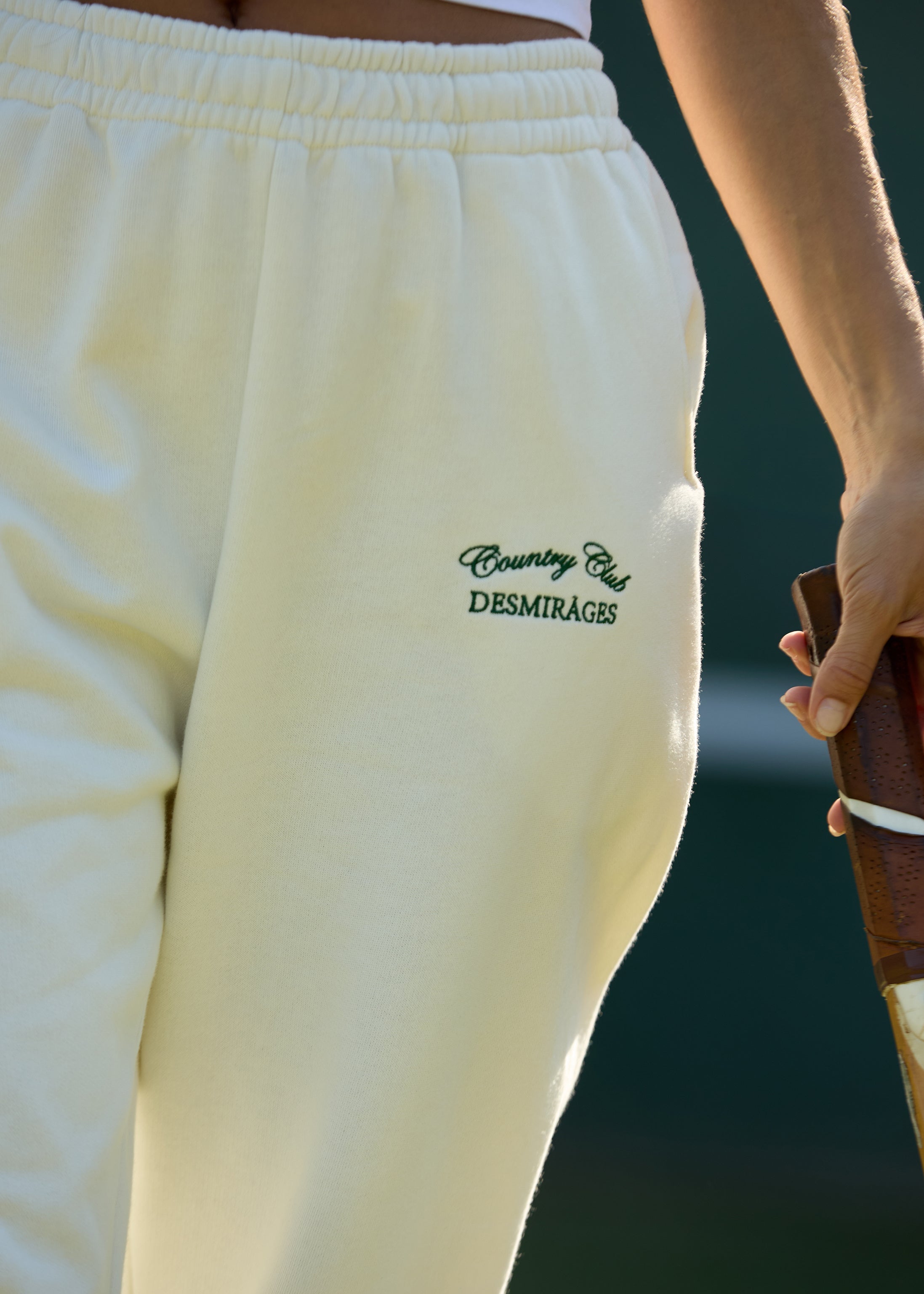 Close-up of the elastic waistband, side pocket and green DESMIRAGES Country Club embroidery on cream sweatpants.